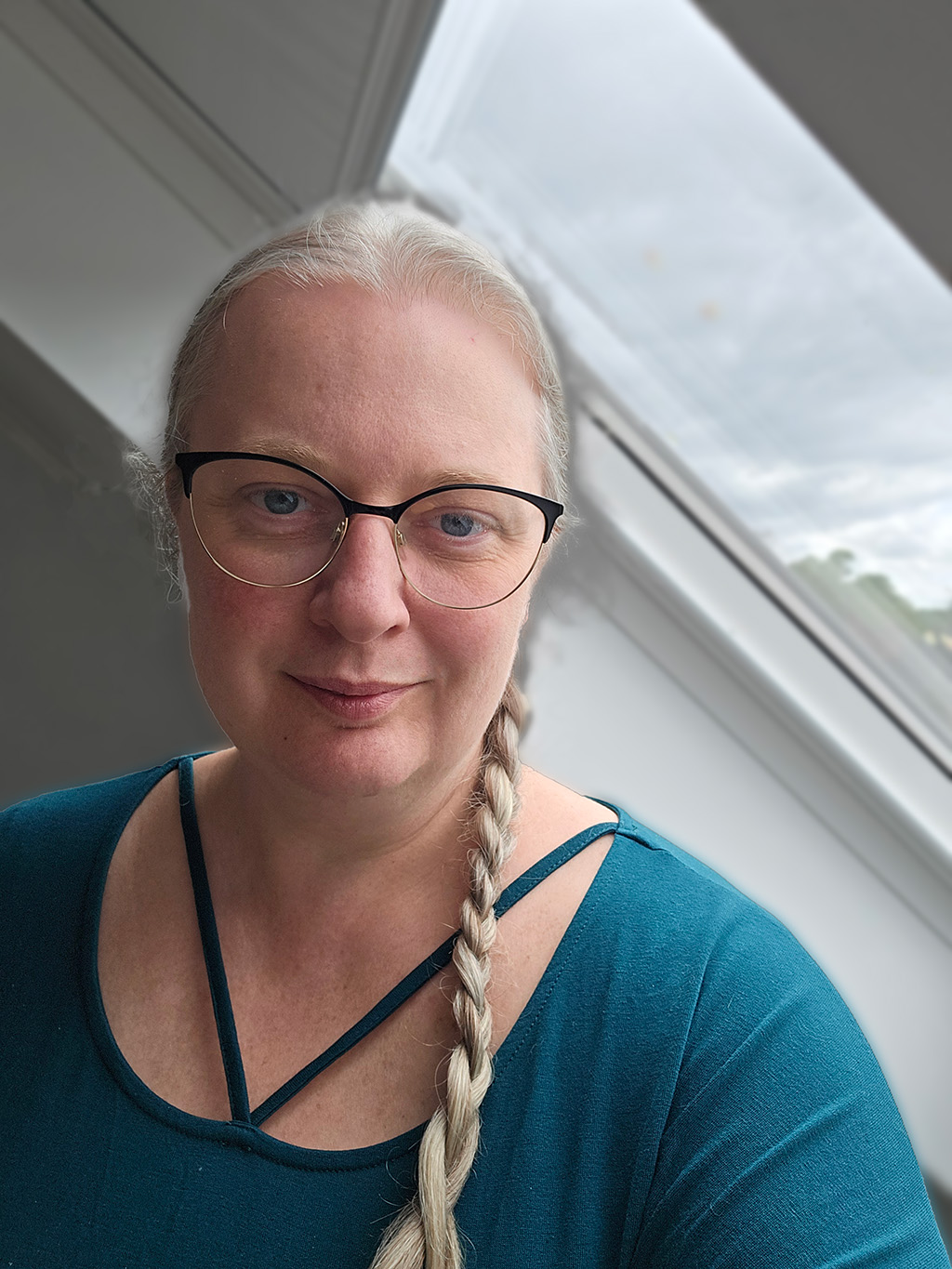 Joanna, smiling by a window, glasses on and hair in a long braid.